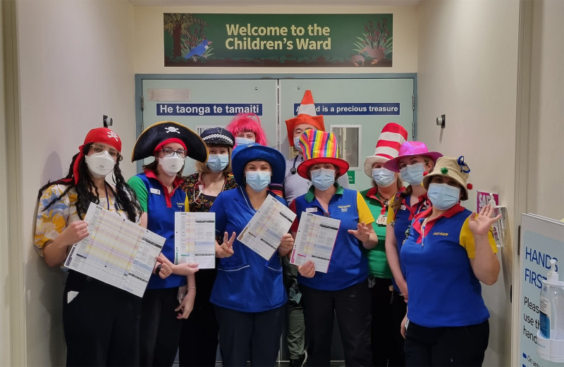 An image of a group of clinicians, dressed up in various funny hats pose in front of the entrance to the Children's Ward holding up PEWS documents.