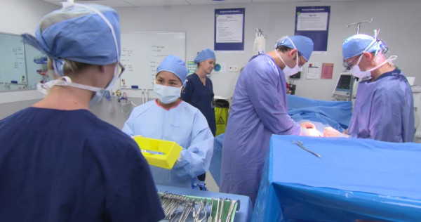 An image of a surgery in progress. There are five clinicians in the room all wearing scrubs and protective equipment. The patient is hidden by a privacy shield.