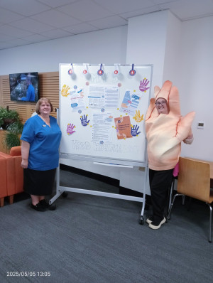 Two people standing next to a whiteboard. One person is in scrubs and the other is dressed in a costume shaped like a hand. The whiteboard has the words 'hand hygiene' written on it and is decorated with information about hand hygiene day.