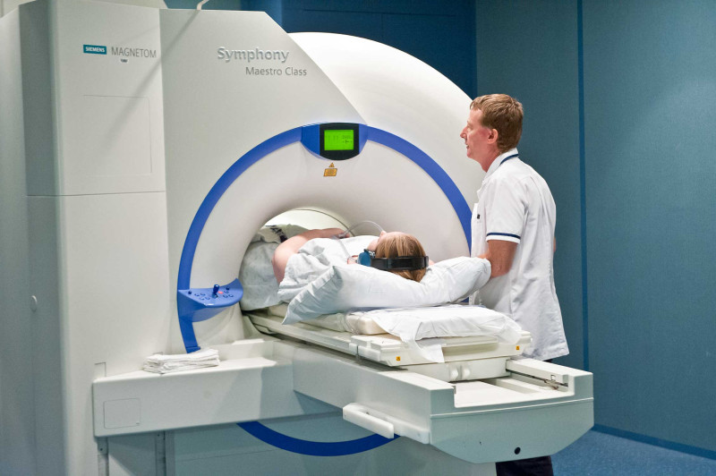 A woman (Jane) lying on her back with her head propped up on pillows, about to go through a tunnel-shaped, scanning machine. A nurse in white scrubs stands next to her for support.  