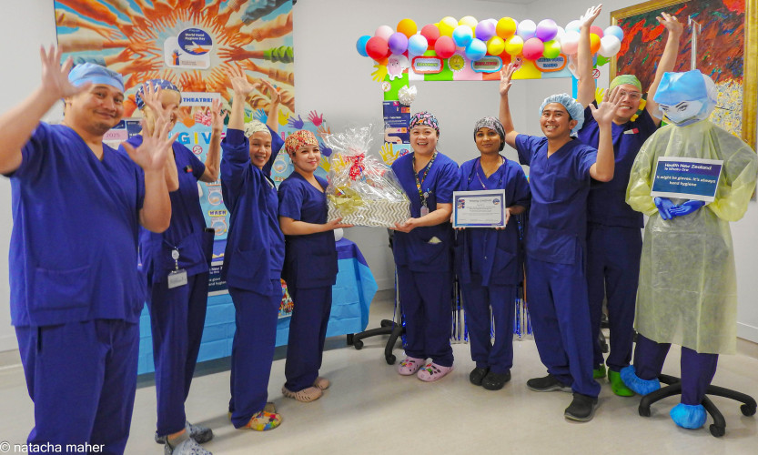 A group of people in scrubs with their hands up in front of balloons and a poster. To the right is a dummy dressed in scrubs and disposable hygiene protective equipment.