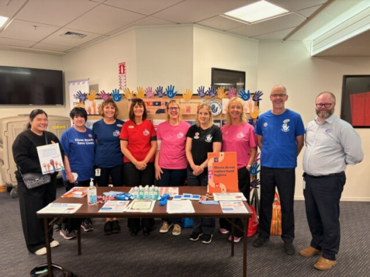 A group of people standing together behind a table that has hand hygiene posters and pamphlets and bottles of hand sanitiser on it.