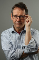 A head and shoulders photo of Jeffrey Braithwaite, who is wearing a white and blue checked shirt and reading glasses. He is smiling and holding the corner of his glasses with one hand while his other arm is crossed over his chest.