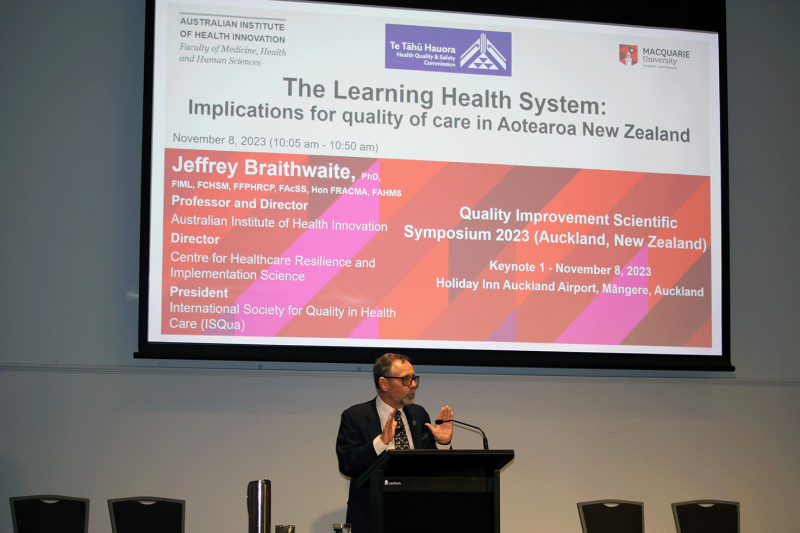 An image of Professor Jeffery Braithwaite presenting on stage. Above his head is a screen projecting his presentation powerpoint titled, 'The Learning Health System: Implications for quality of care in Aotearoa New Zealand'.