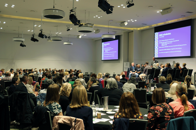 An image of a conference room with groups of people sitting around tables. In the middle right of the frame is a stage with chairs and people sitting facing one another. The screens above them show a powerpoint with Te Tāhū Hauora branding titled 'mihi whakatau'.