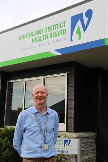 Joe stands in a blue shirt in front of a building with the sign Northland District Health Board visible above his head.