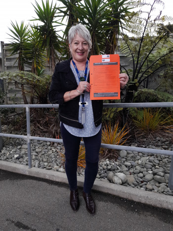 Paula stands in front of a garden, holding the new orange envelope related to their project