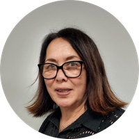 Carol has straight shoulder length brown hair. She is wearing large black framed glasses and a black shirt with patterned cutouts