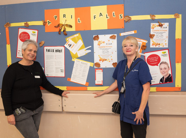 An image of two clinicians standing in front of a wall display with various posters about falls. The title of the wall display is 'April Falls'.